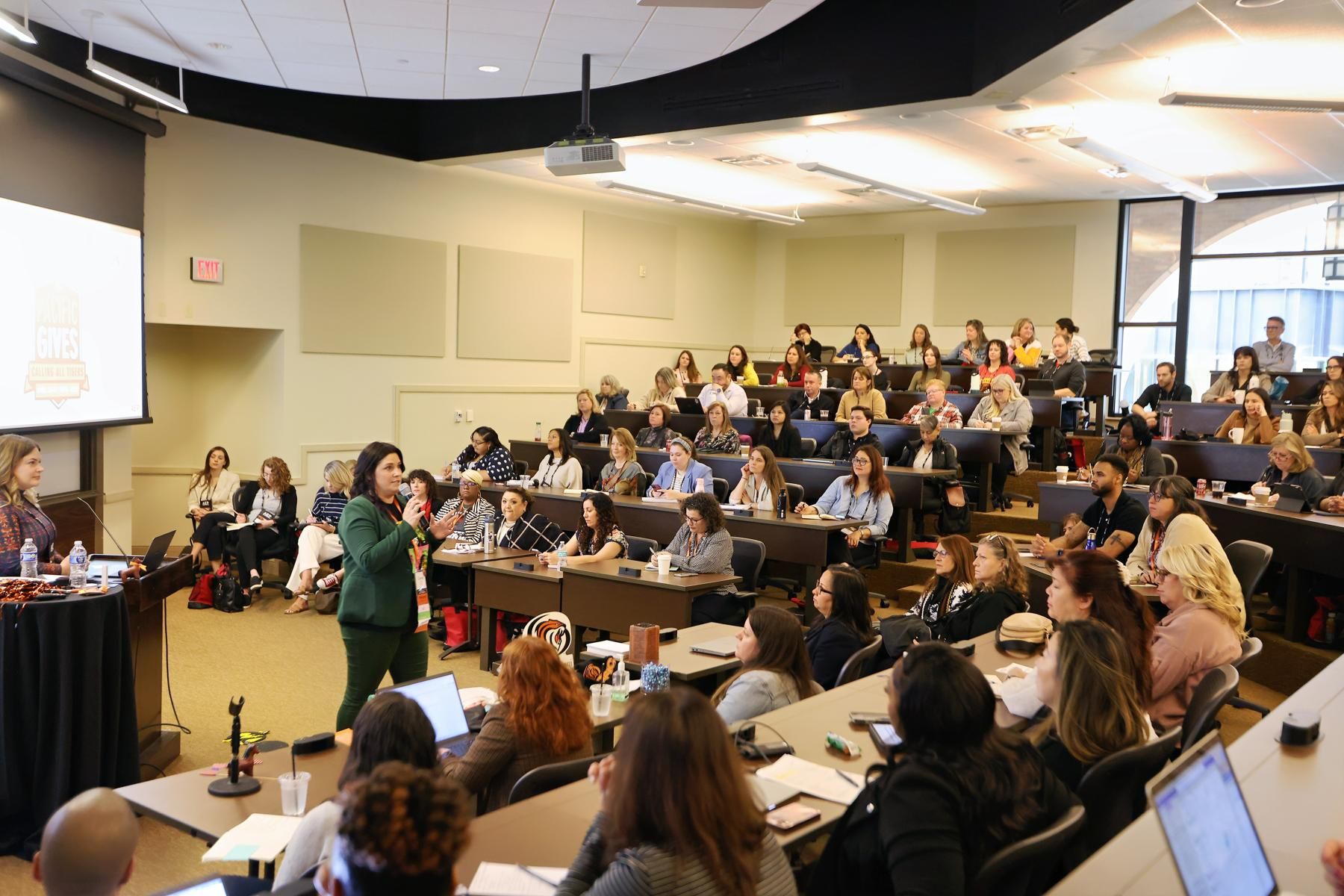 A speaker presents to a full classroom of attendees seated in tiered rows during a professional conference or workshop. The audience is engaged, taking notes and listening attentively, while a presentation slide is displayed on a screen at the front of the room.