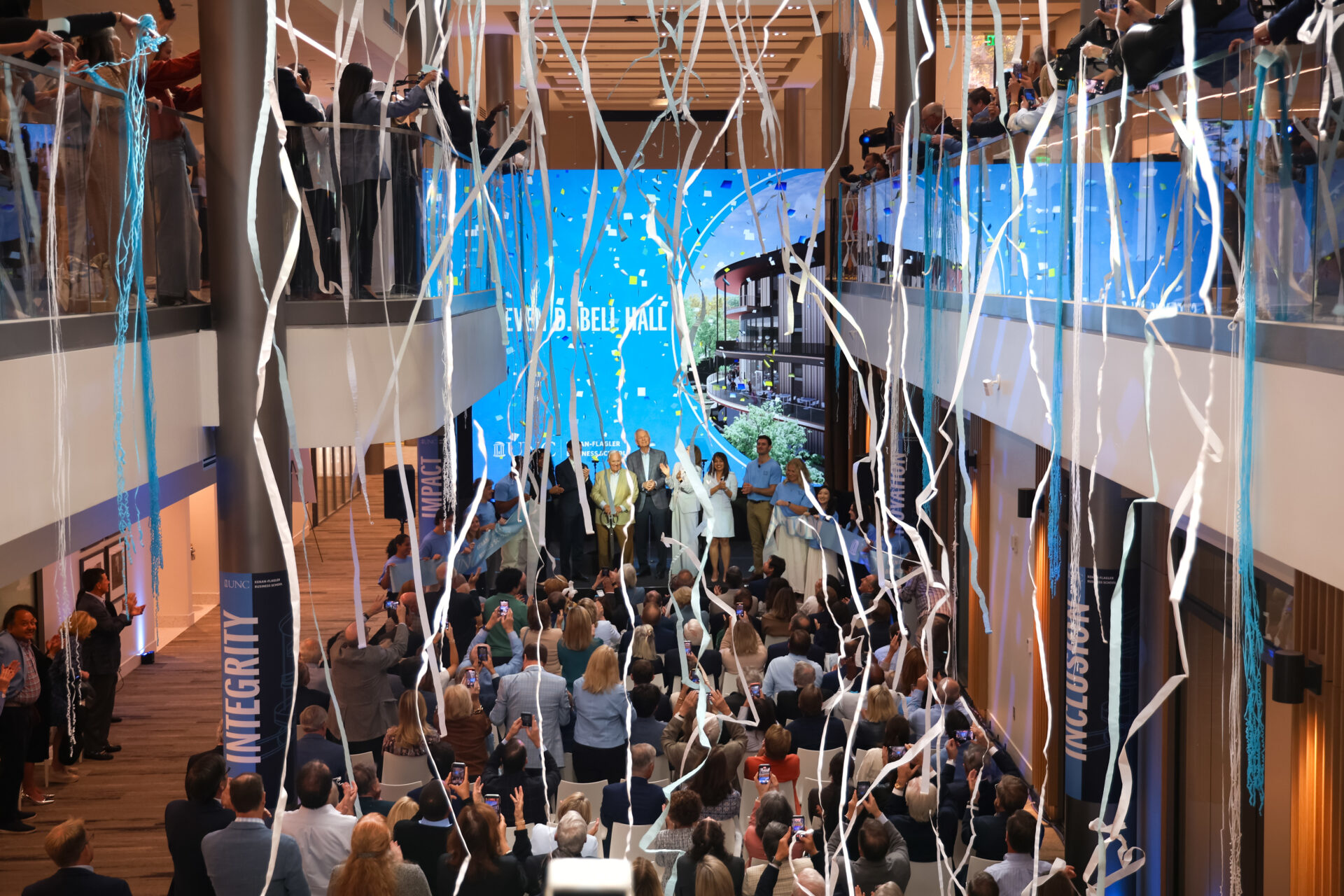 Grand opening ceremony in multi-level atrium with speakers on central stage, large crowd on multiple floors, and white and blue streamers hanging from ceiling.