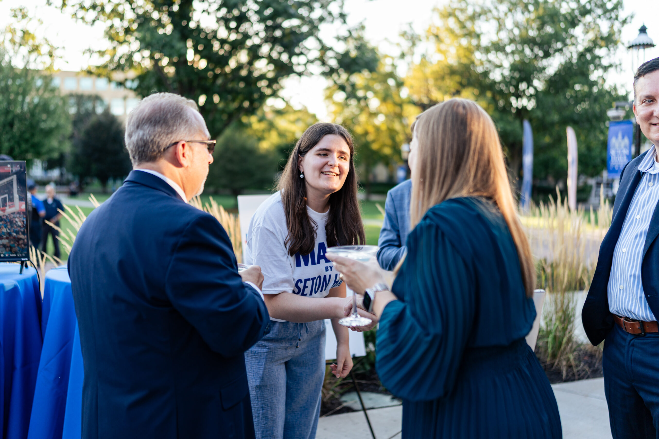 A young woman wearing a white t-shirt with "SETON Hall" text smiles while conversing with four adults at an outdoor event. The group stands in a campus setting with large shade trees, manicured landscaping, and a blue-clothed table visible on the left. The woman is holding a cocktail. The adults are dressed in business casual attire—navy blazers and striped shirts. Golden afternoon sunlight filters through the trees, and a brick building with a cupola is visible in the background.