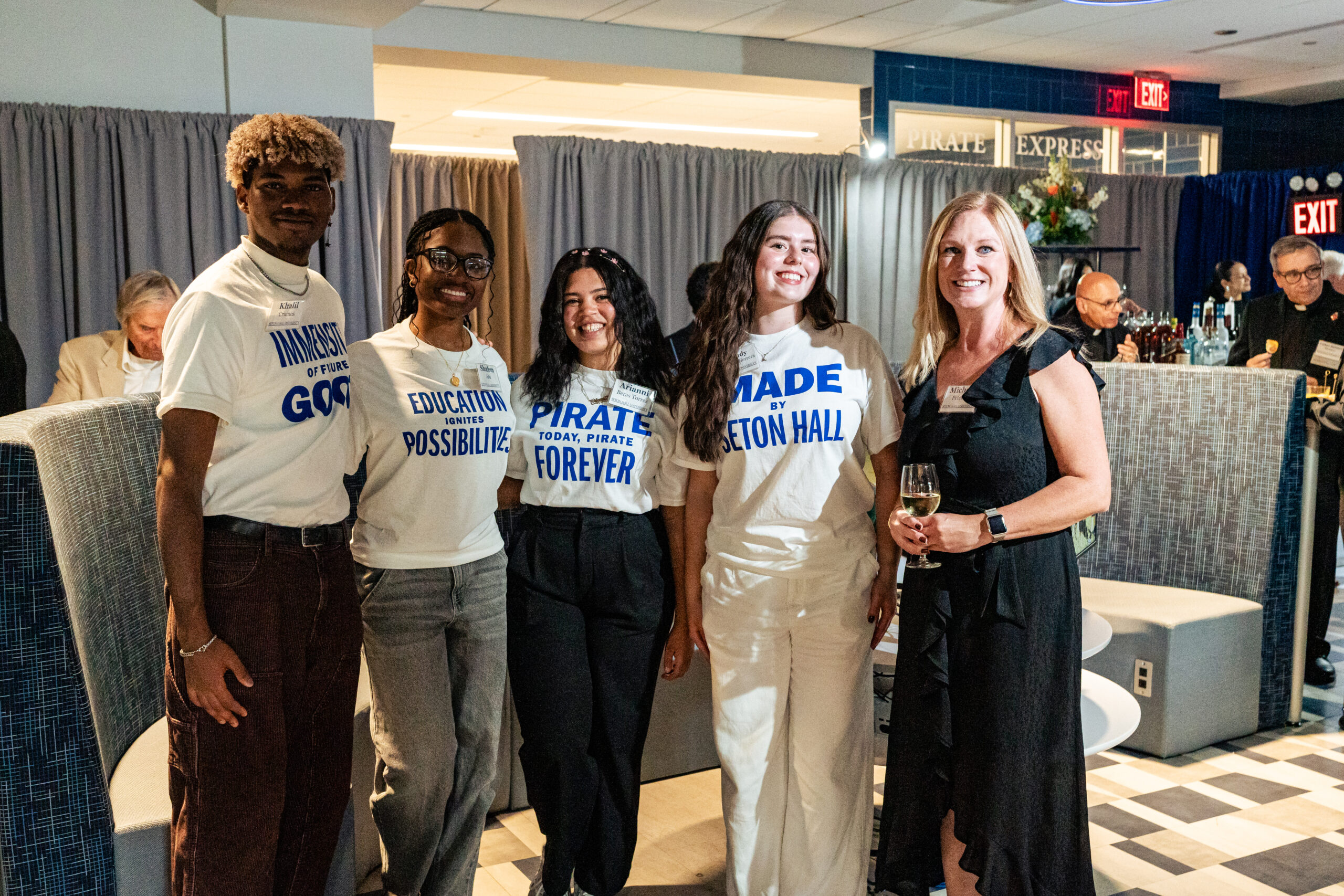 A group of five people stand together smiling at an indoor event. Four young adults wear white T-shirts with bold blue Seton Hall–themed messages, and the fifth person, an adult woman in a black dress, holds a glass of white wine. They are posing in a lounge-style area with soft seating, curtains, and a bar in the background.