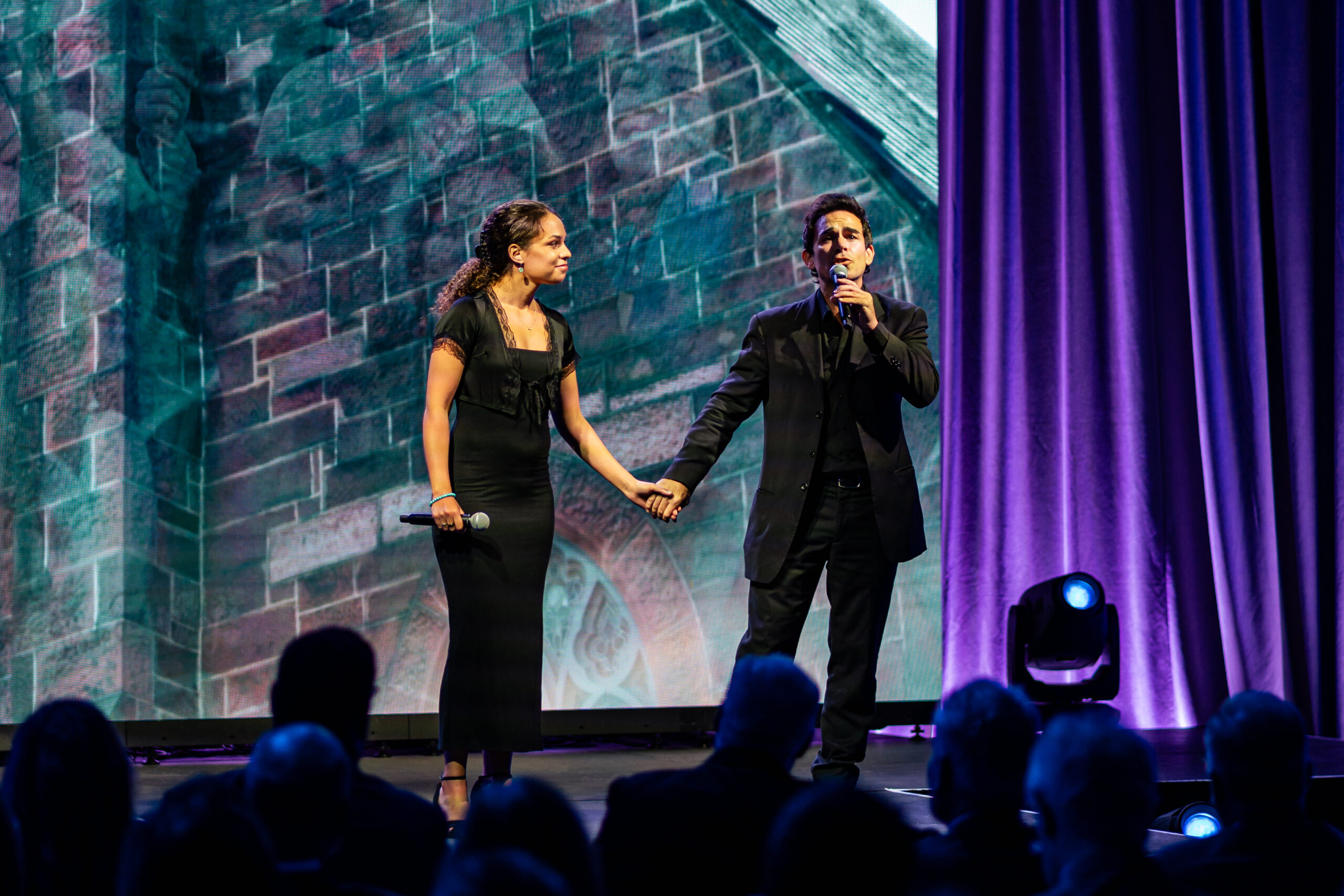 Two performers stand on stage holding hands during a university campaign launch event program. The woman on the left wears a black sleeveless dress and holds a microphone, while the man on the right wears a black suit and shirt and speaks into a microphone. Behind them is a large LED screen displaying a teal and pink brick wall pattern. Purple stage lighting illuminates the backdrop and curtains on either side. An audience is visible in silhouette in the foreground, with some audience members holding up phones to record the event.