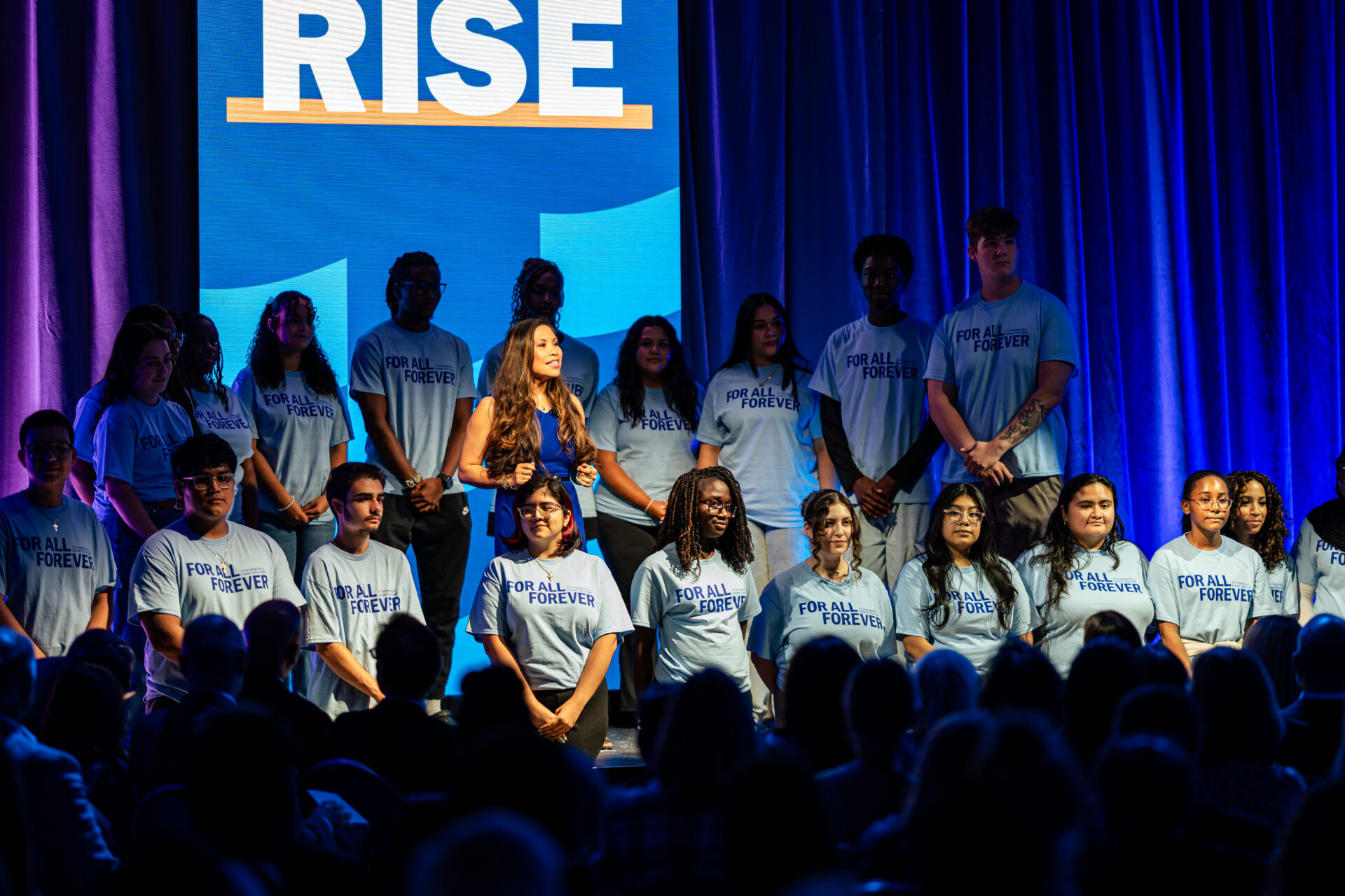 A diverse group of young people wearing matching gray t-shirts with "FOR ALL FOREVER" printed in blue text stand on stage under dramatic blue and purple lighting. They are arranged in multiple rows in front of a large LED screen displaying the word "RISE" in white letters with an orange underline. The stage is lit with vibrant purple and blue uplighting, and an audience is visible in the foreground. The setting is a university campaign launch event with an inspirational theme.
