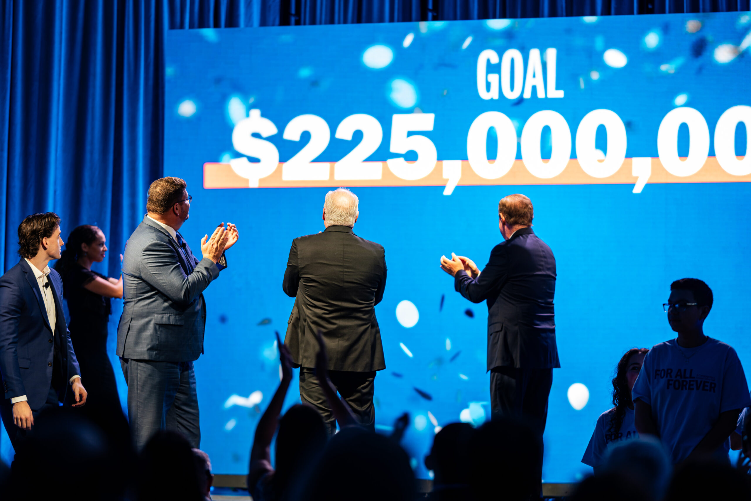 A group of people on stage applaud as a large screen behind them reveals a campaign fundraising goal of $225,000,000. Blue lighting and falling confetti create a celebratory atmosphere, while audience members cheer from the foreground.