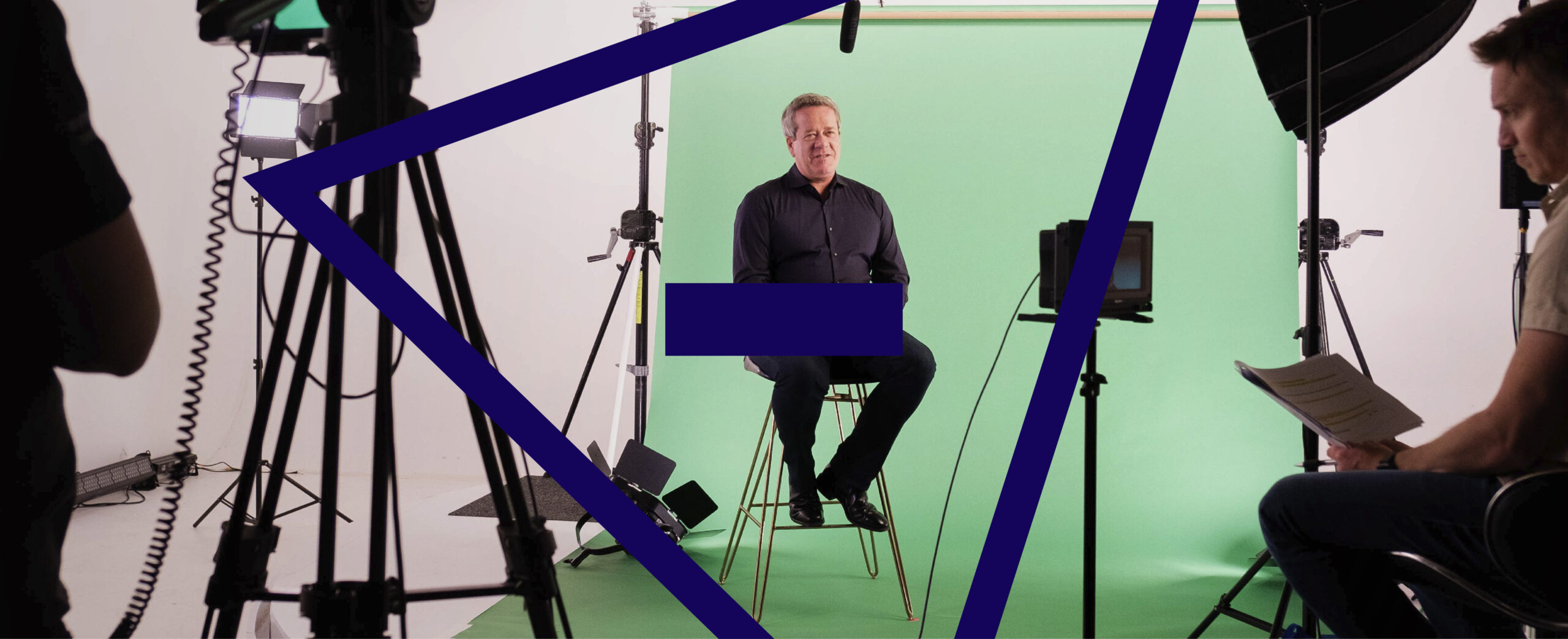 A man sits on a stool with a greenscreen behind him on a camera set.