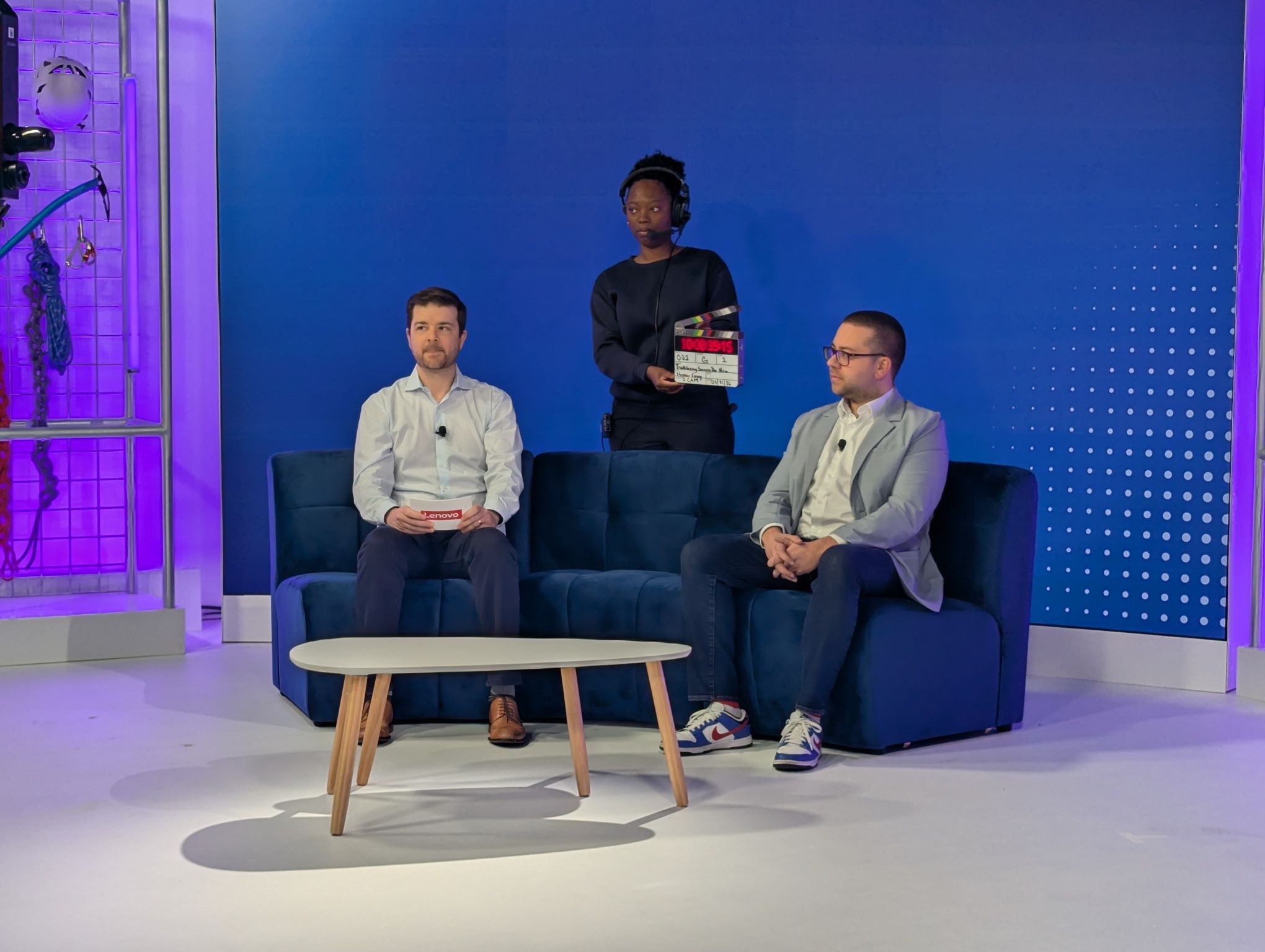 Two presenters seated on a navy blue studio sofa with a Lenovo-branded set, while a production crew member holds a clapperboard behind them ahead of a live bro…Two presenters seated on a navy blue studio sofa with a Lenovo-branded set, while a production crew member holds a clapperboard behind them ahead of a live broadcast taping.