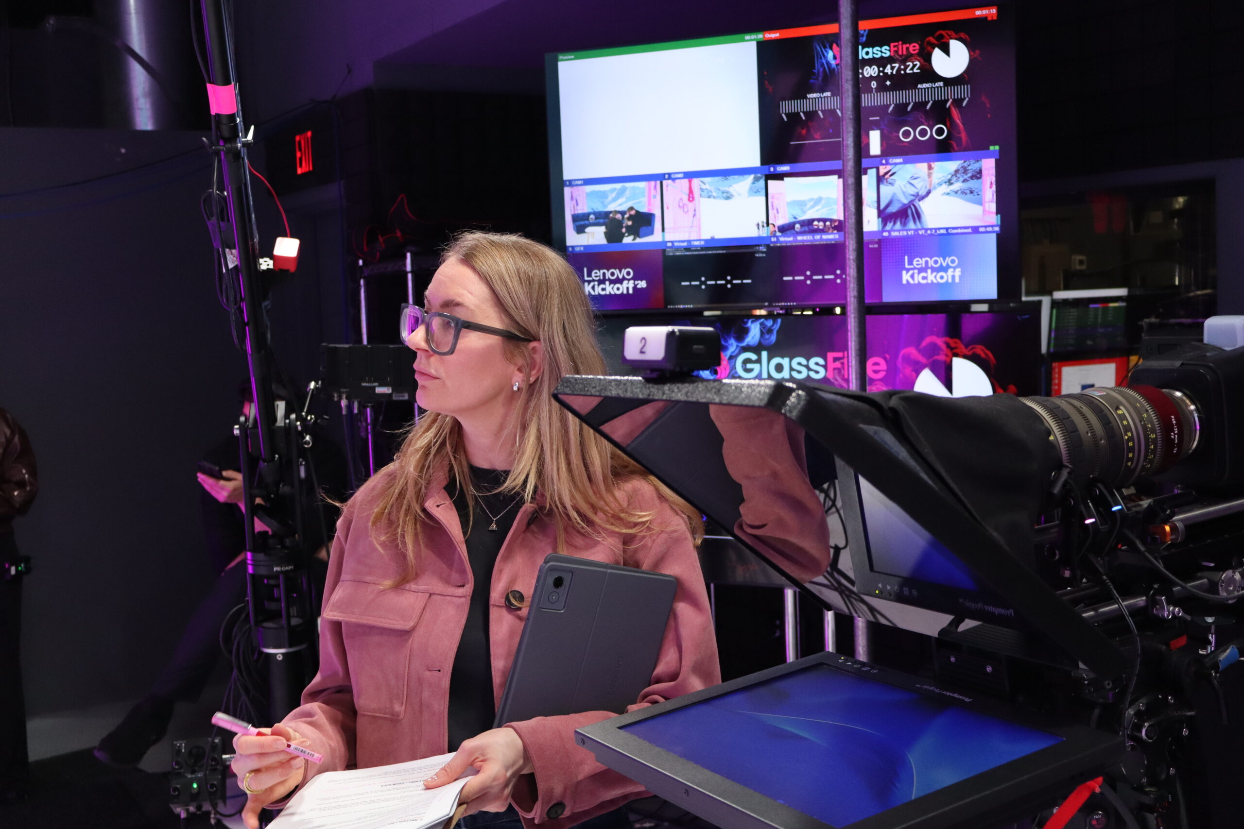A production manager monitors the live broadcast from behind the cameras at TSEC Studios, holding a tablet and run-of-show notes with Lenovo Kickoff '26 broadcast screens visible in the background.
