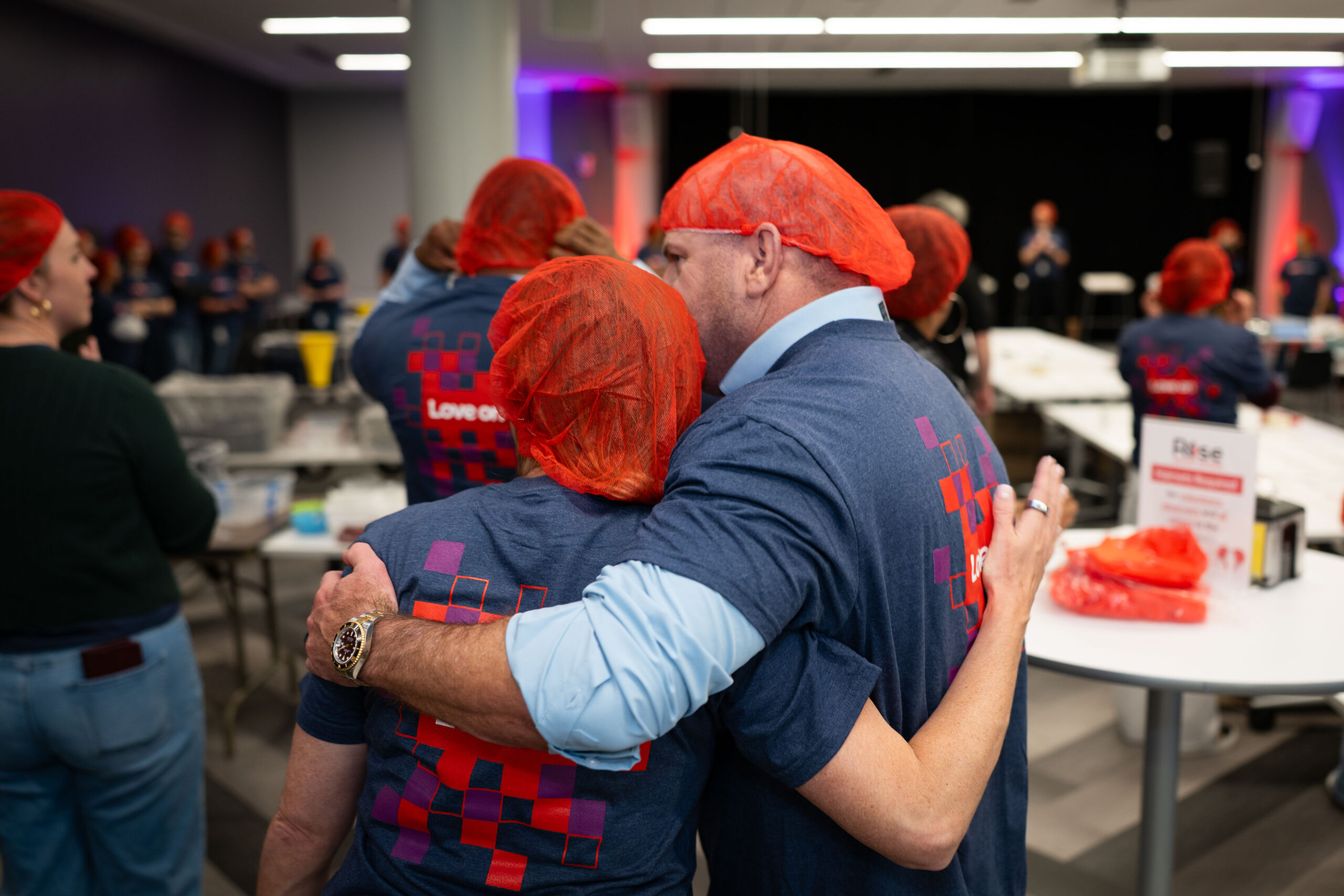 Lenovo employees wearing hairnets and branded t-shirts embrace during the Rise Against Hunger CSR activity, where attendees packed meals for communities in need as part of the Lenovo Kickoff '26 hybrid event.