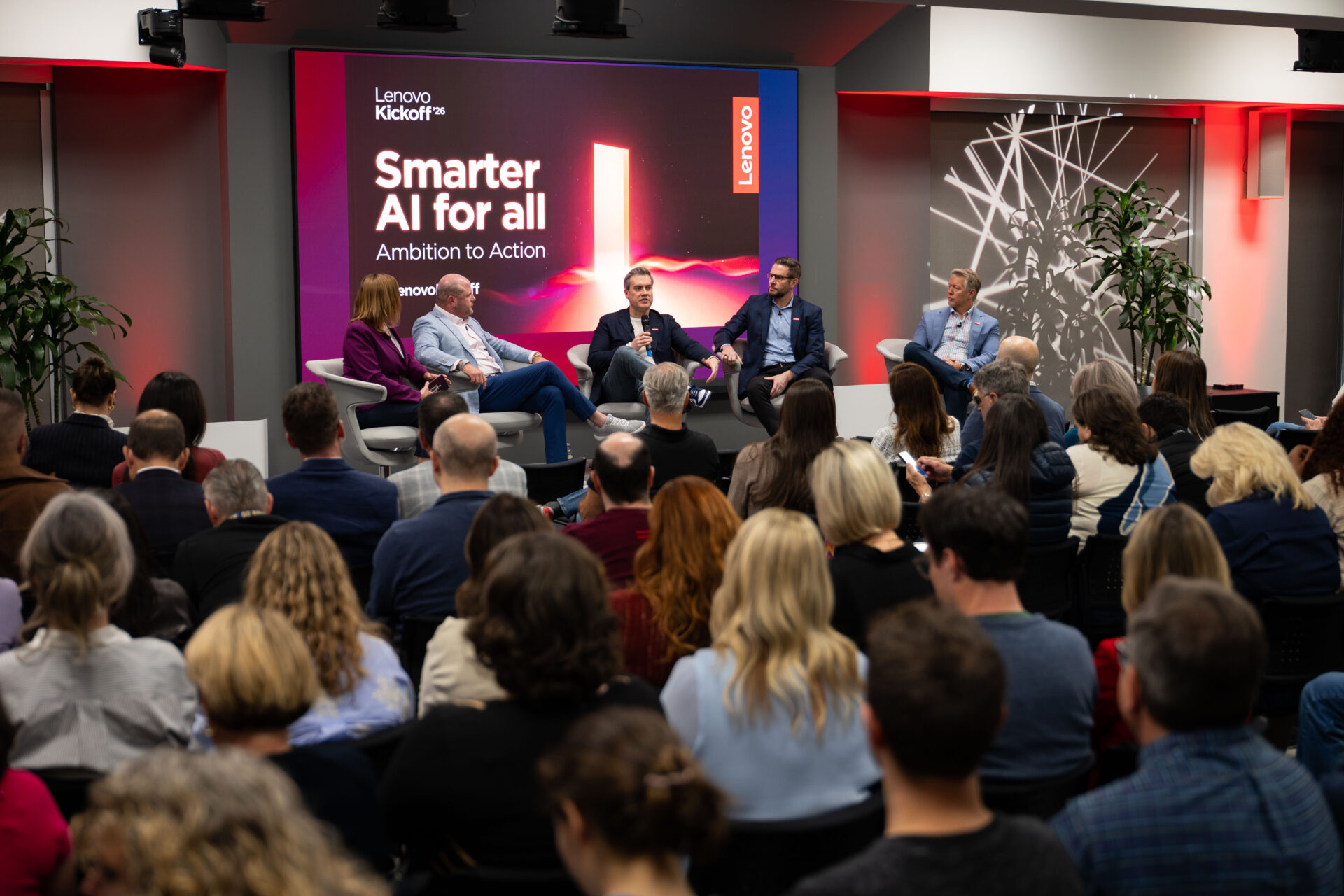 A panel discussion at Lenovo Kickoff '26, themed "Smarter AI for all – Ambition to Action." Five panelists sit in modern chairs on a stage in front of a large branded screen displaying the Lenovo logo and event title. A full audience viewed from behind fills the foreground, seated in rows facing the stage. The venue has warm accent lighting with red and purple tones.