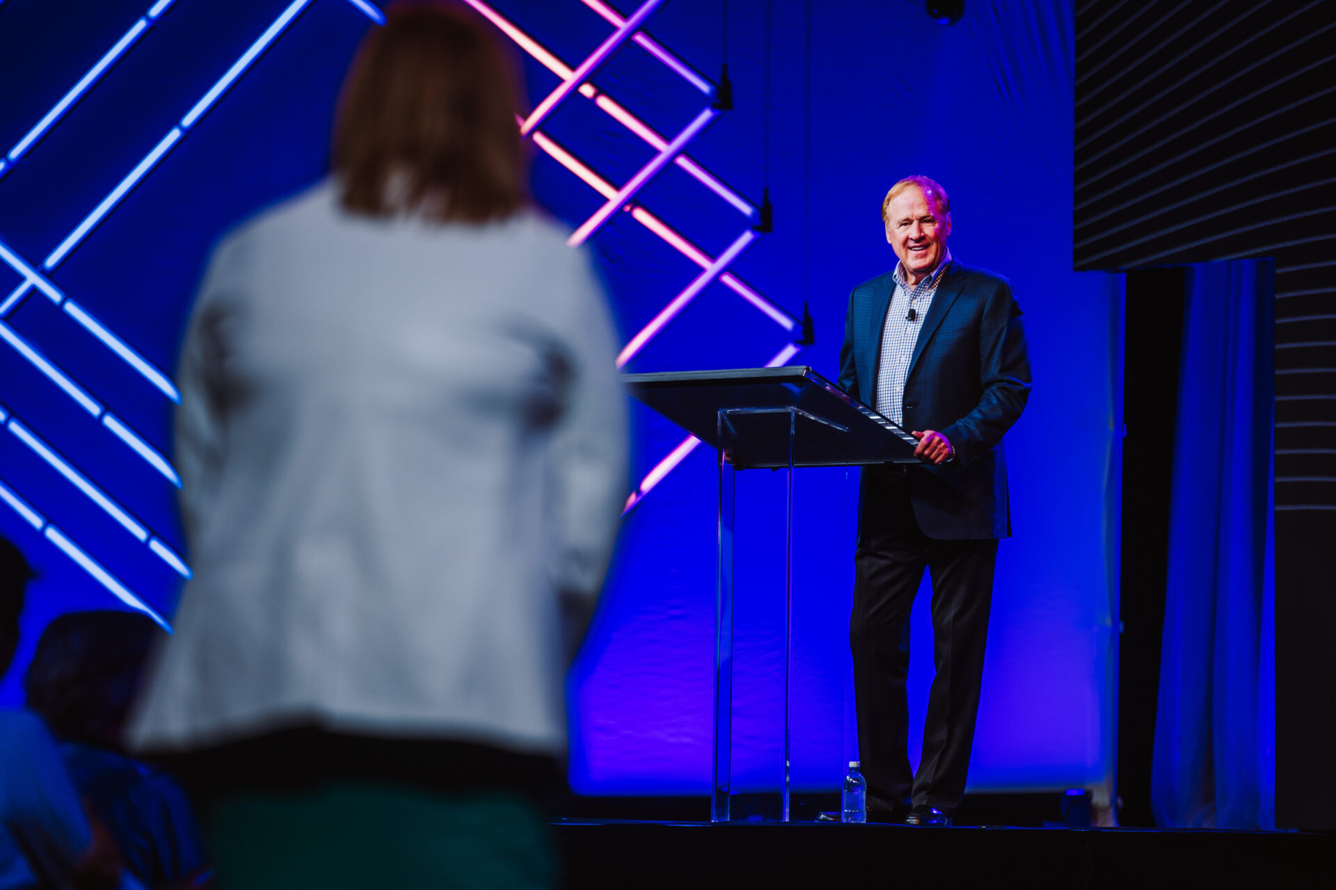 A confident speaker stands at a glass podium on a professional stage with striking neon geometric patterns in blue and magenta. An attendee in light clothing is visible in soft focus in the foreground, suggesting an engaged audience at a modern conference or corporate event.
