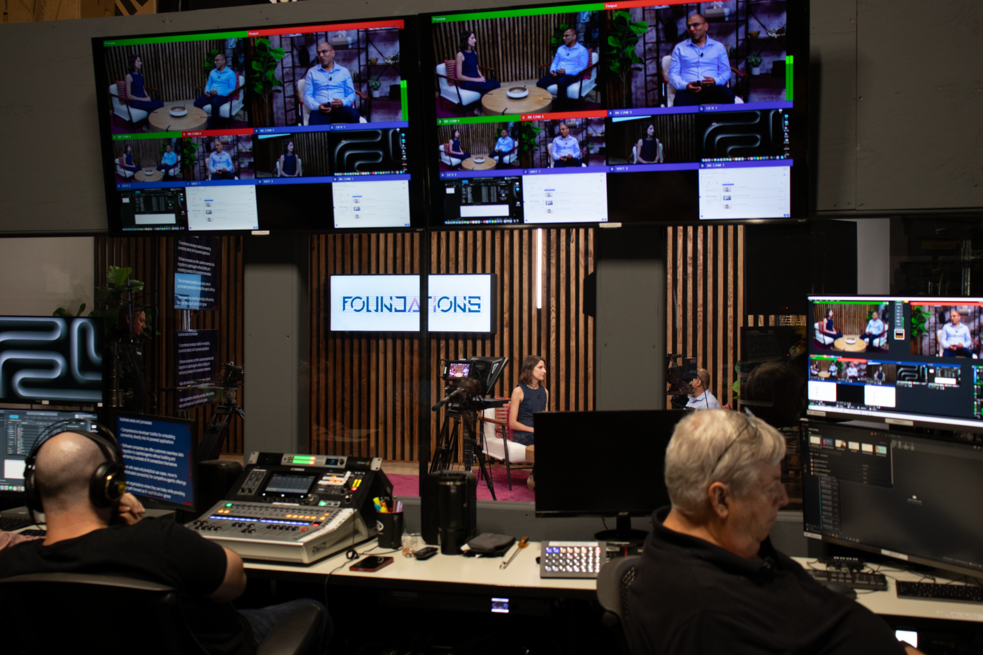 Broadcast control room displaying a professional virtual event streaming setup, with two operators at desks managing production equipment and monitors showing multiple video feeds of panelists on large displays above. The "FOUNDATION" branding is visible on a wooden-slat backdrop, and camera and audio equipment are positioned for live event coverage.