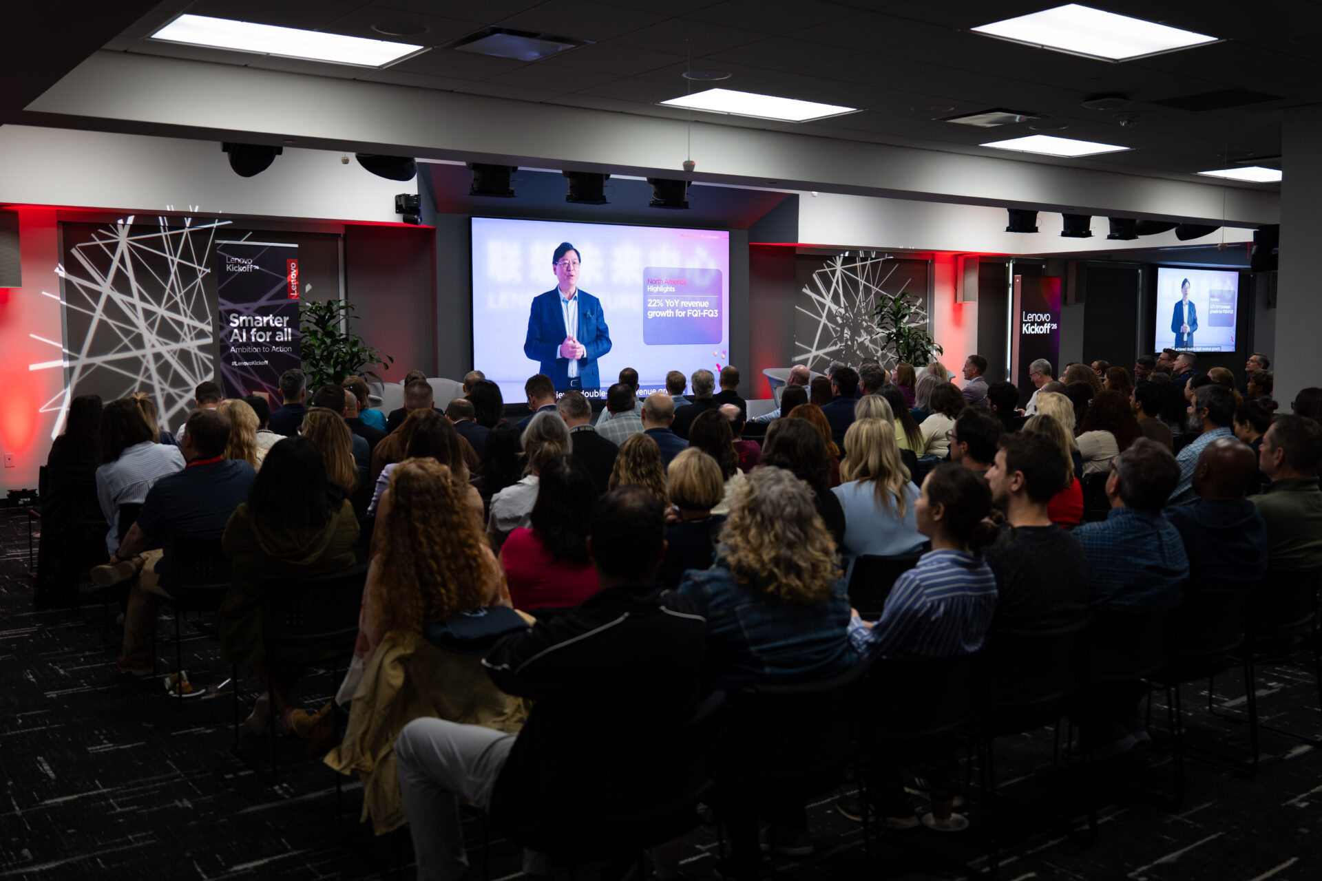 Large group of corporate hybrid event attendees watching a keynote presentation on a projection screen at a Lenovo kickoff event, with red and black branded walls framing the modern venue.