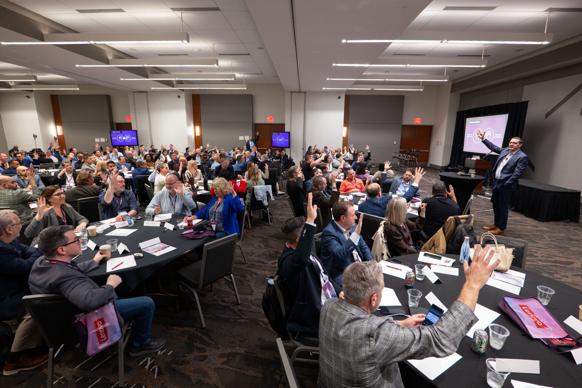 A crowded conference room with a speaker presenting at the front and many audience members seated at round tables with hands raised in participation.